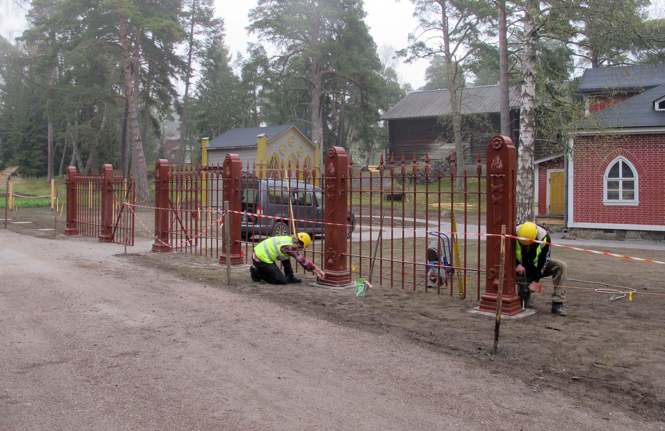 Kaksi työasuista henkilöä työskentelemässä rauta-aidan vieressä. Aitaosa on harva ja siro, mutta sen pylväät ovat jämäkät ja koristeelliset. Taustalla näkyy huvimaja ja muita rakennuksia.