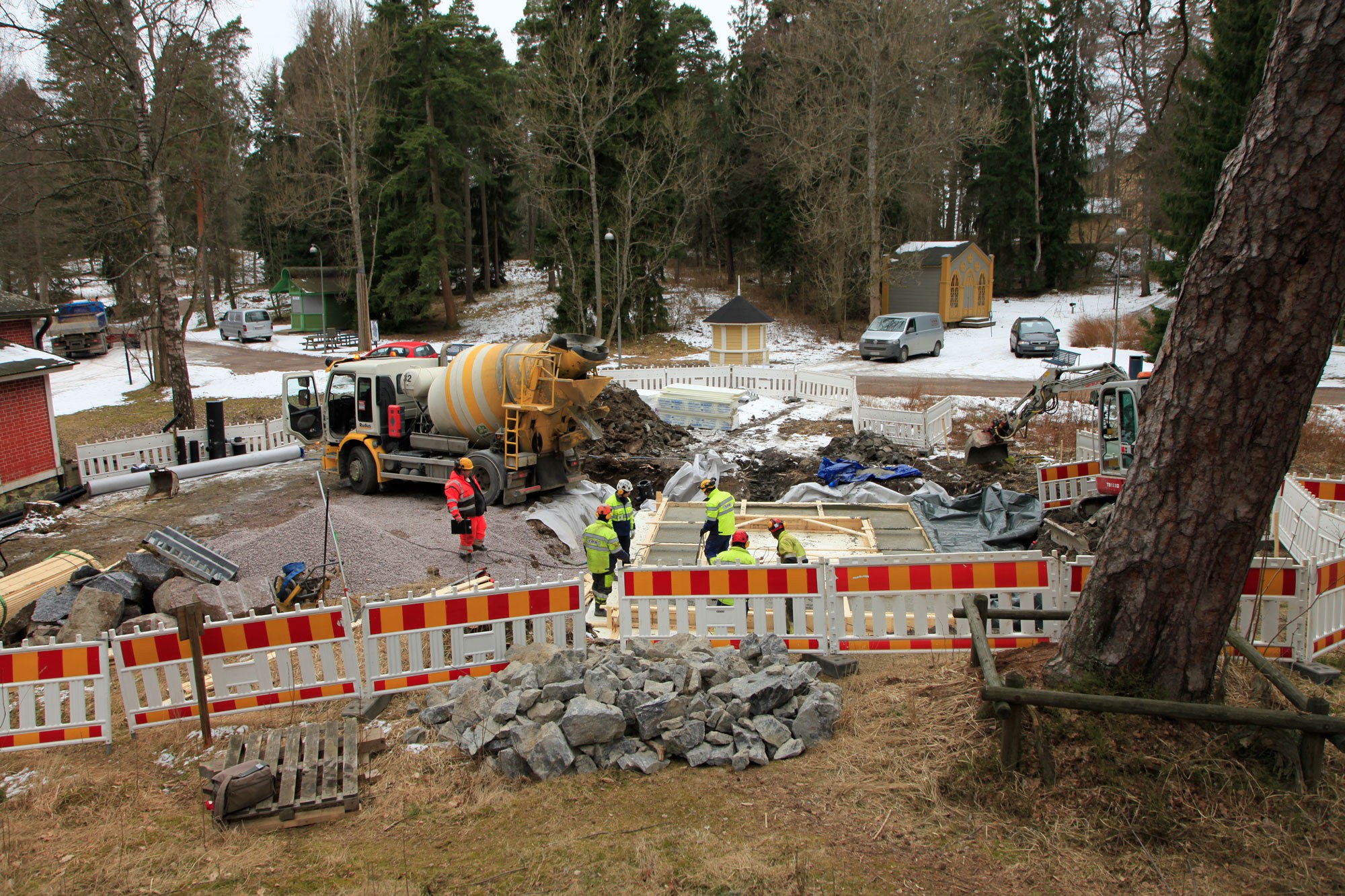 Työmaa-aitaa, sementtirekka, huomioasuisia ihmisiä ja perustusten puinen kehys.