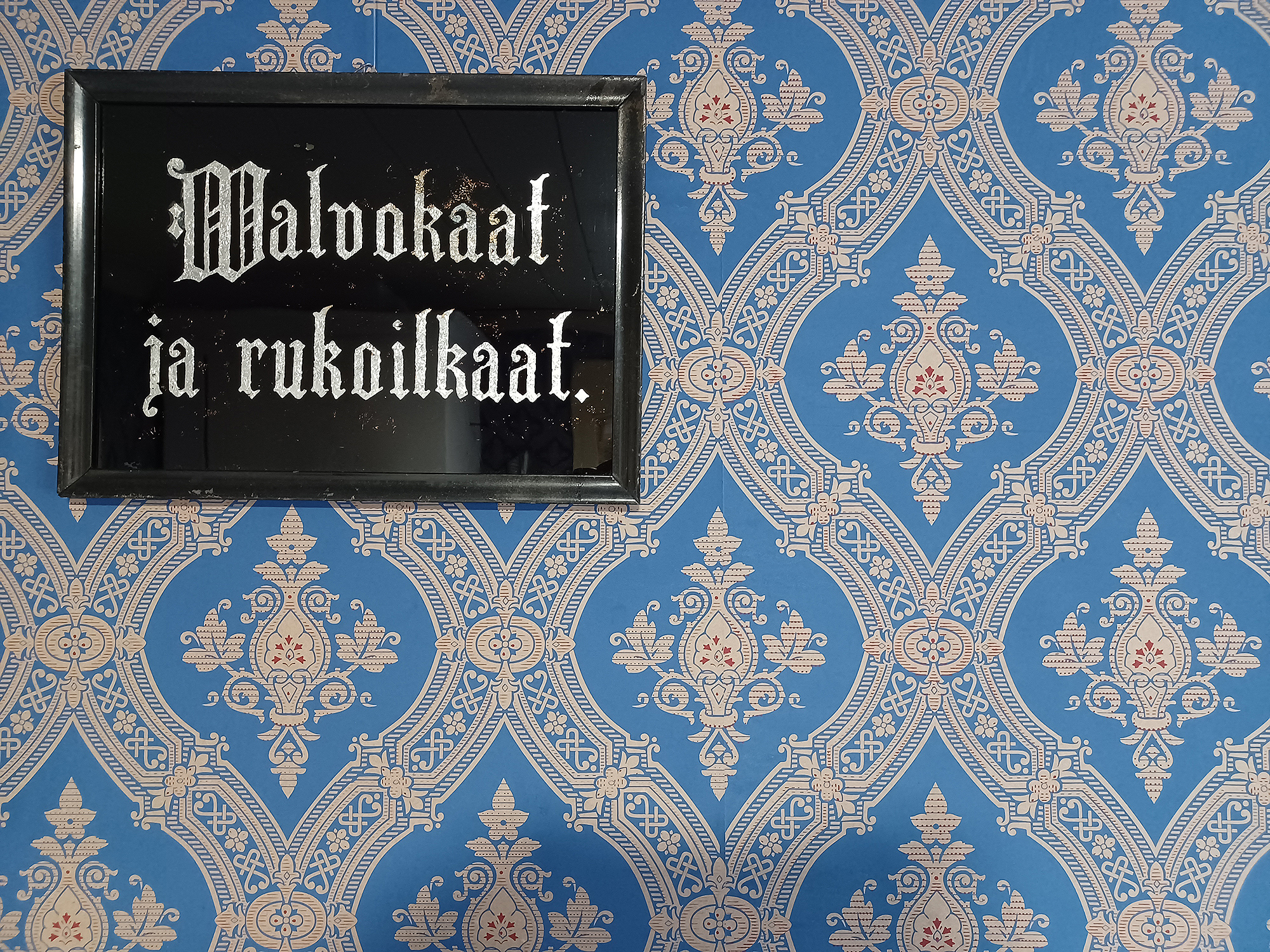 Seinällä on taulu, jossa lukee Walvokaat ja rukoilkaat! Seinässä on tiheäkuvioinen, koristeellinen tapettipinta.
