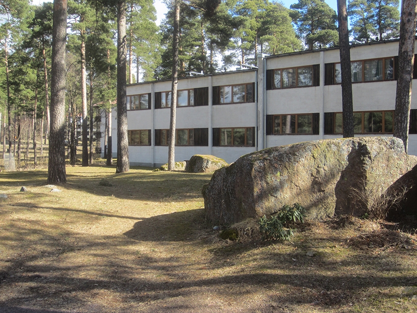 Valkea kaksikerroksinen asuinkerrostalo mäntyjen lomassa.