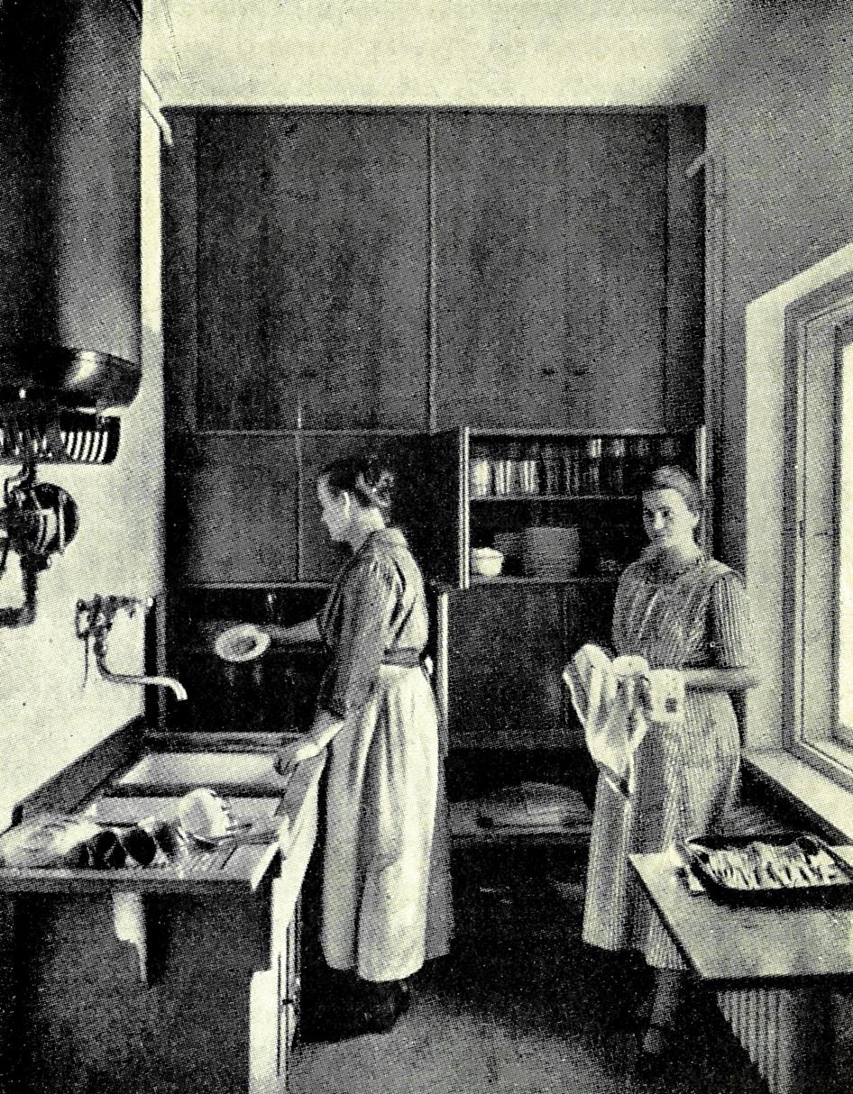 Vanha valokuva keittiöstä, jossa kaksi henkilöä työskentelee tiskialtaan ja kaappien äärellä.